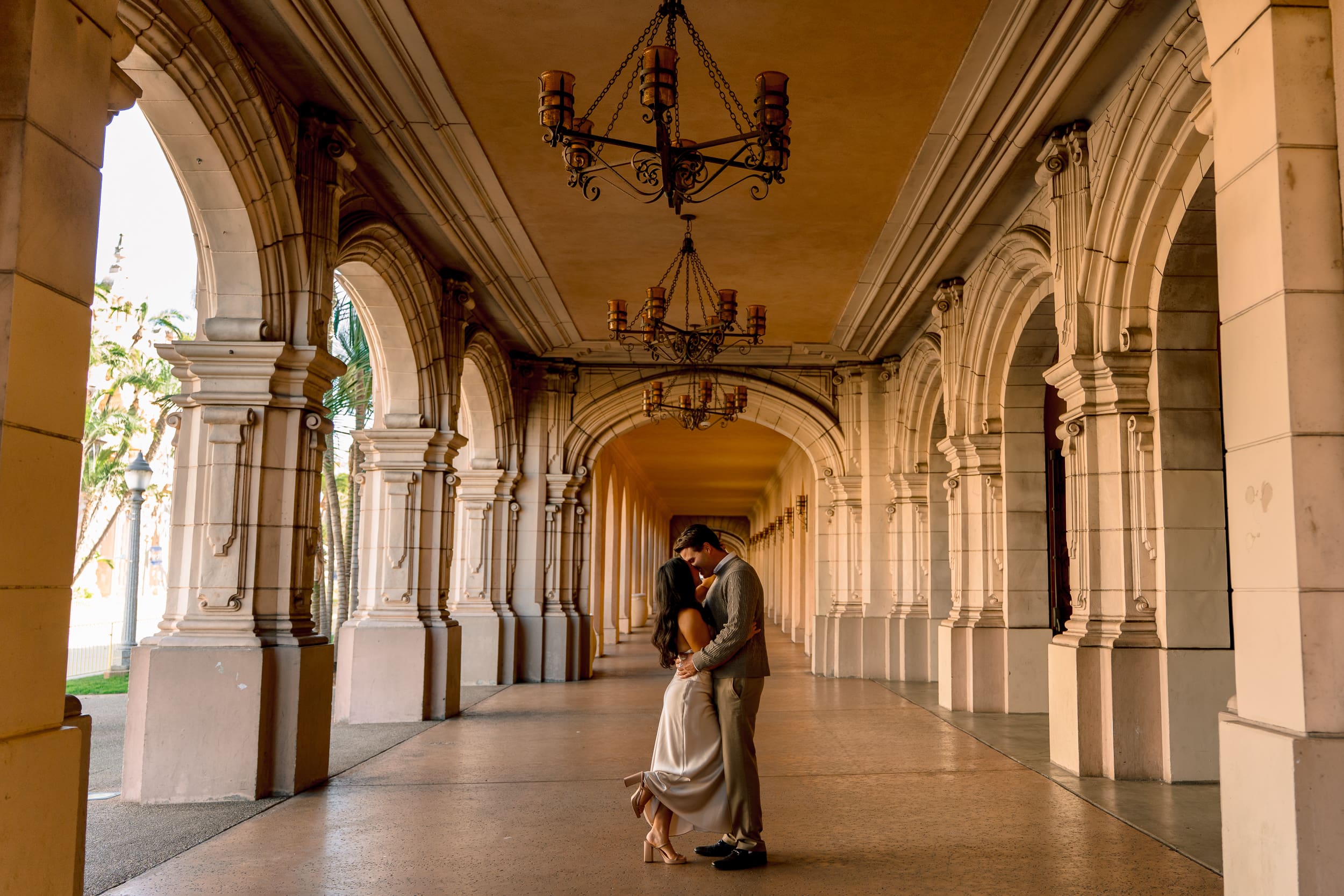 Wide-angle editorial engagement portrait under the iconic Balboa Park arches, featuring warm golden hour light and romantic posing.