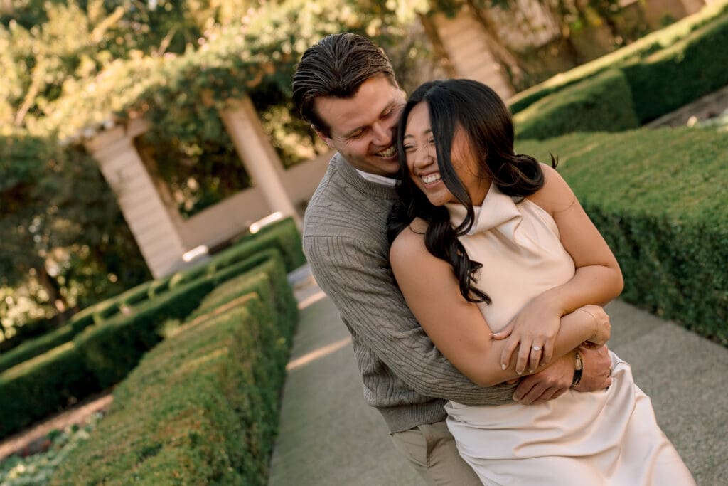 Candid photo of a couple laughing while he hugs her from behind in the formal garden at Balboa Park during their engagement session.