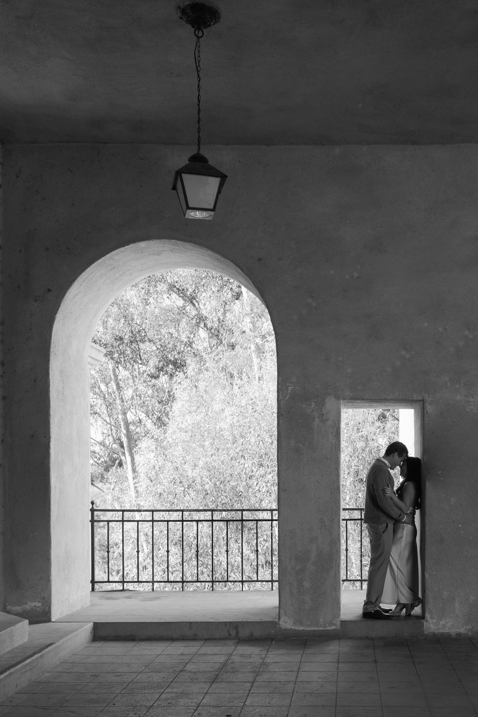 Black and white editorial engagement photo of a couple standing in an arched doorway at Balboa Park, framed by dramatic shadows and natural light.