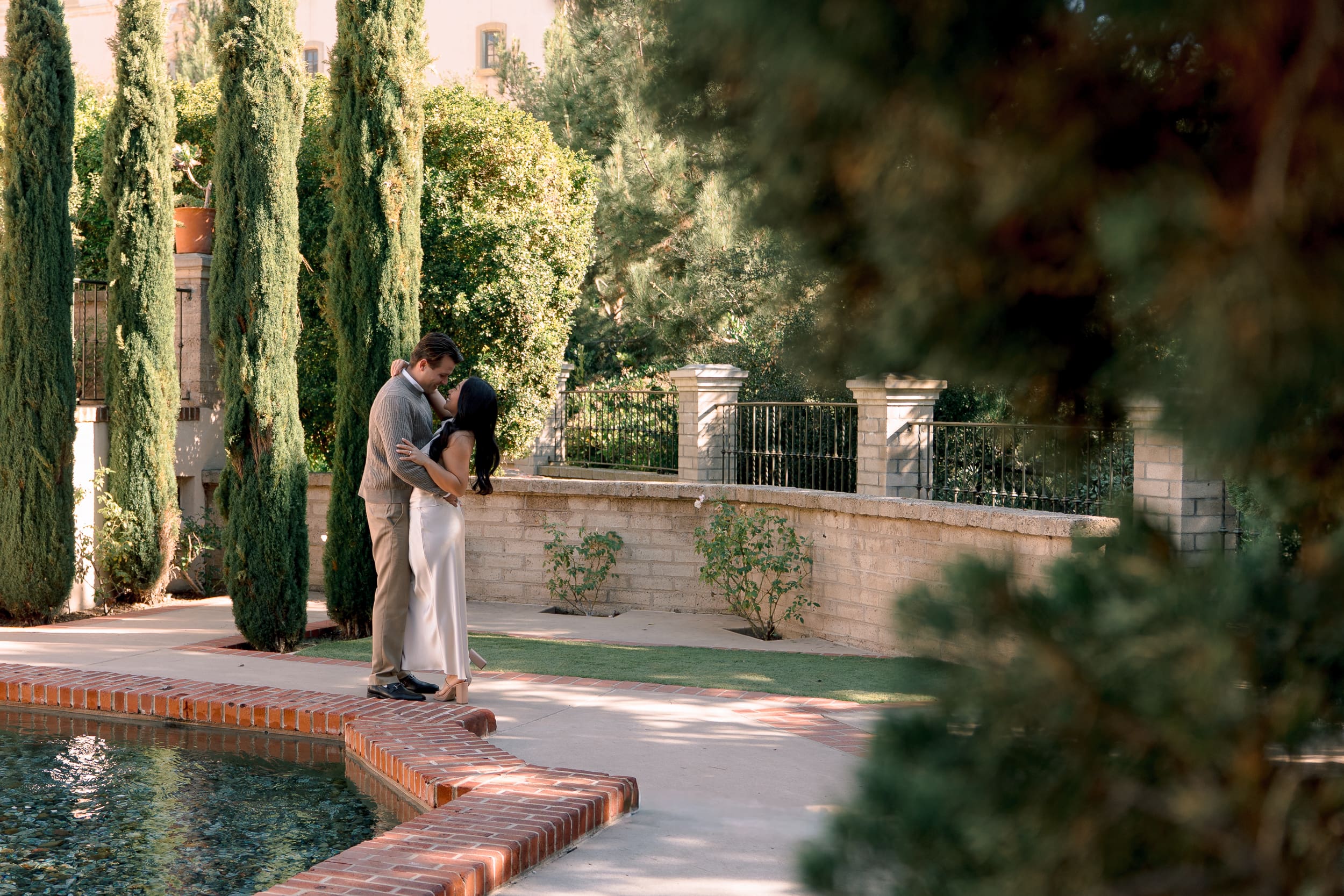 Romantic Balboa Park engagement photo of a couple embracing near tall cypress trees, captured by San Diego engagement photographer Ro Banner during golden hour
