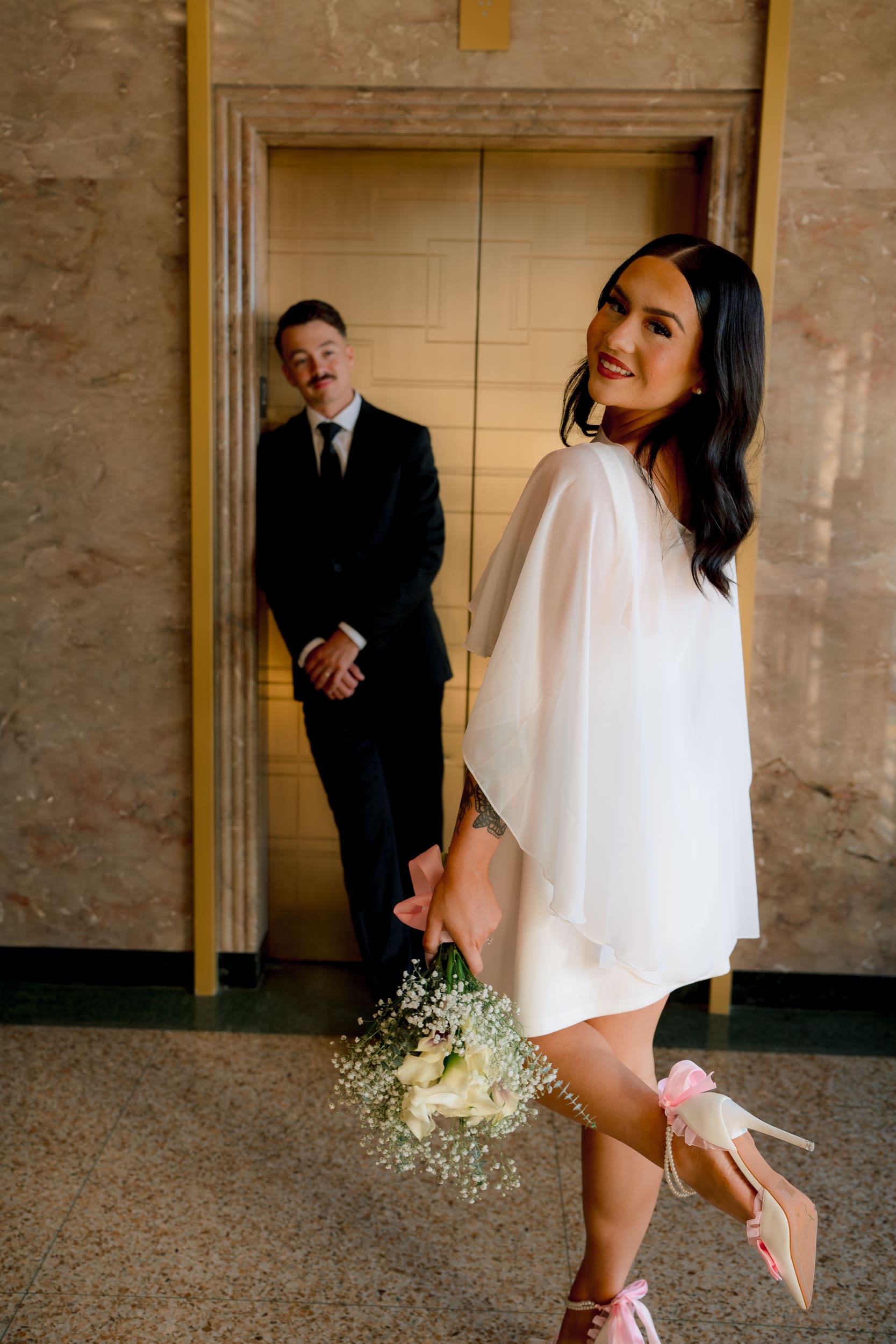Bride looking back while holding her bouquet in the courthouse hallway during a San Diego courthouse wedding, with the groom standing behind her, photographed in a soft editorial style