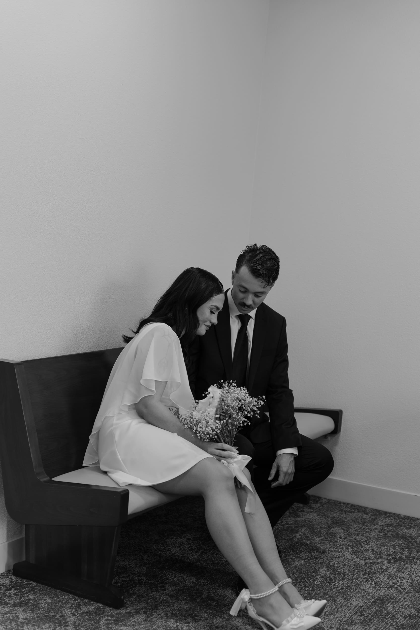 Bride and groom sitting closely together on a courthouse bench during their San Diego elopement, captured in a quiet candid black-and-white documentary style